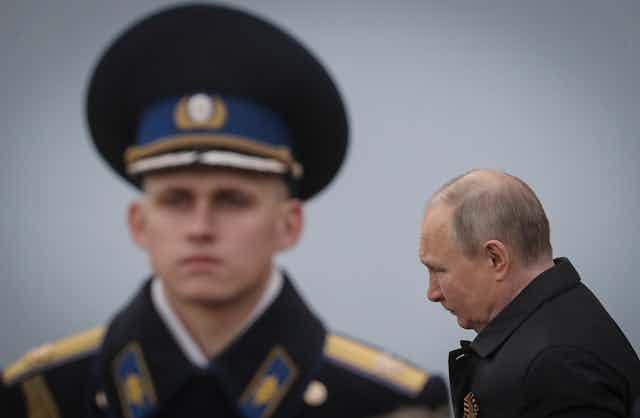 Vladimir Putin in an overcoat with a young military officer in the background.