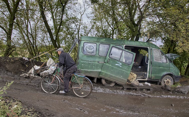 A man rides a bicycle past a damaged army vehicle with the Z symbol.