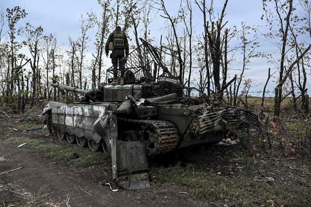 A Ukrainian soldier in fatigues is seen standing atop an abandoned and destroyed Russian tank.