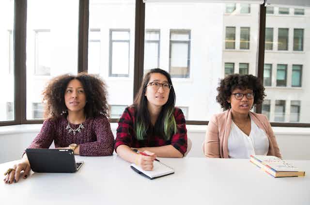Three women: a Black woman, an Asian woman and a Black woman sit together in work conference room with notebooks and laptops. They look confused. 