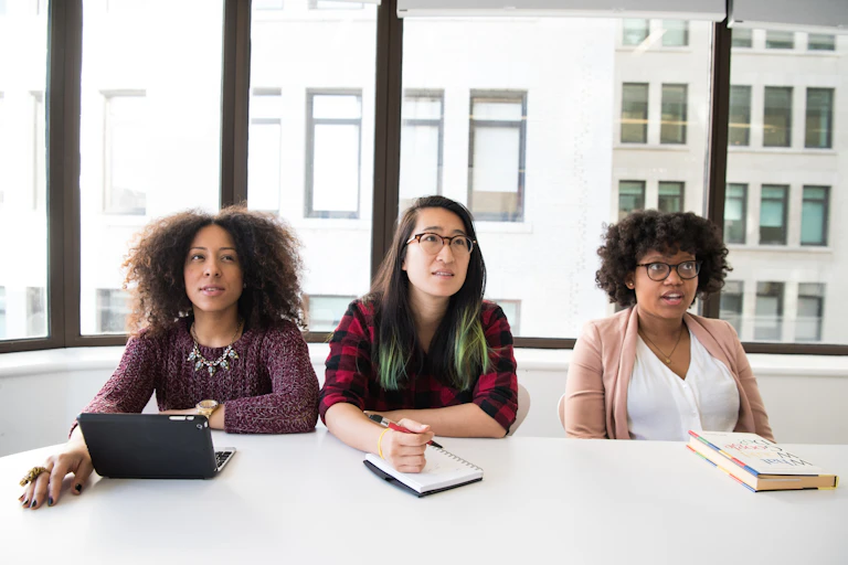 Three women: a Black woman, an Asian woman and a Black woman sit together in work conference room with notebooks and laptops. They look confused.