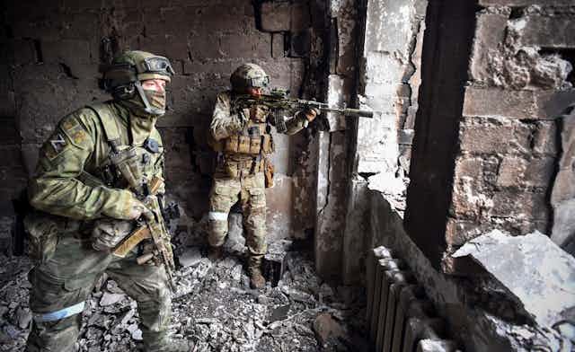Two male soldiers in camouflage are seen holding guns in a. bombed out looking building. One points their gun out of an opening. 