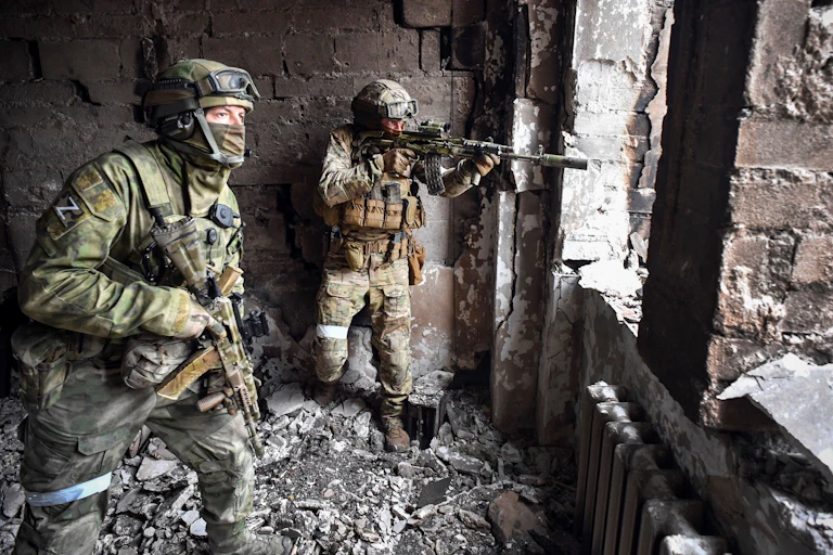 Two male soldiers in camouflage are seen holding guns in a. bombed out looking building. One points their gun out of an opening.