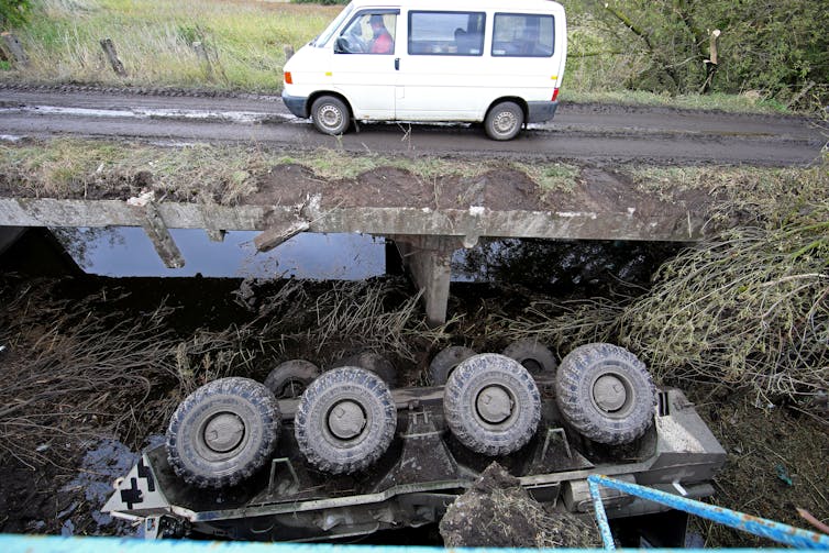 A white van drives over a dirt road and bridge.  Under it a military vehicle is turned upside down, with its four wheels pointing up.