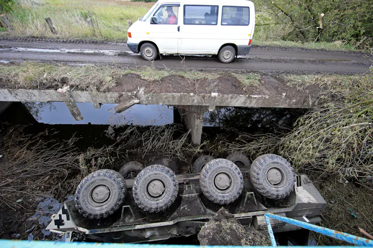 A white van drives over a dirt road and bridge. Under it a military vehicle is turned upside down, with its four wheels pointing up.