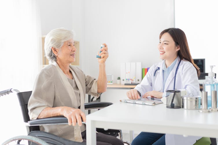 Une jeune femme en blouse et stéthoscope derrière un bureau observe une femme plus âgée qui utilise un inhalateur