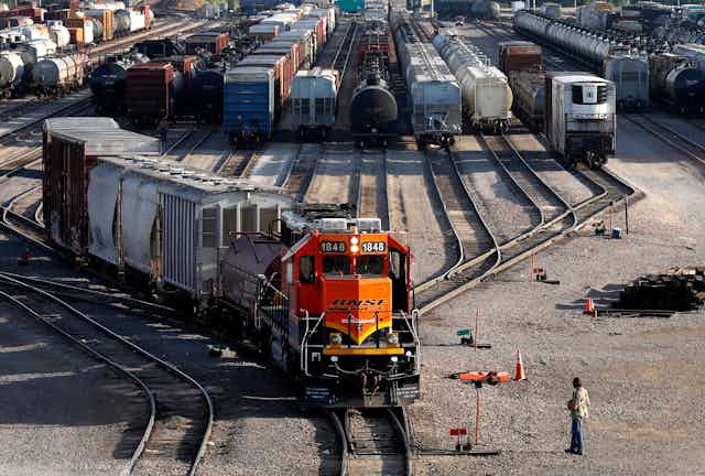 An orange BNSF train engine heads forward with many other train cars in the background and a worker watches