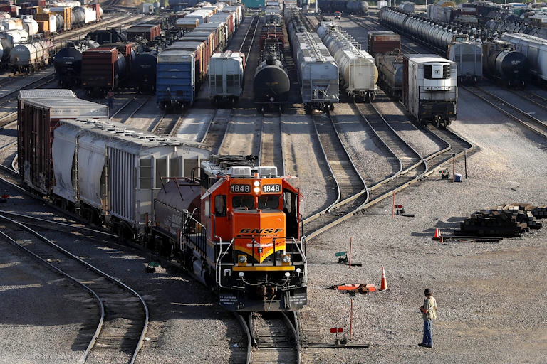 An orange BNSF train engine heads forward with many other train cars in the background and a worker watches