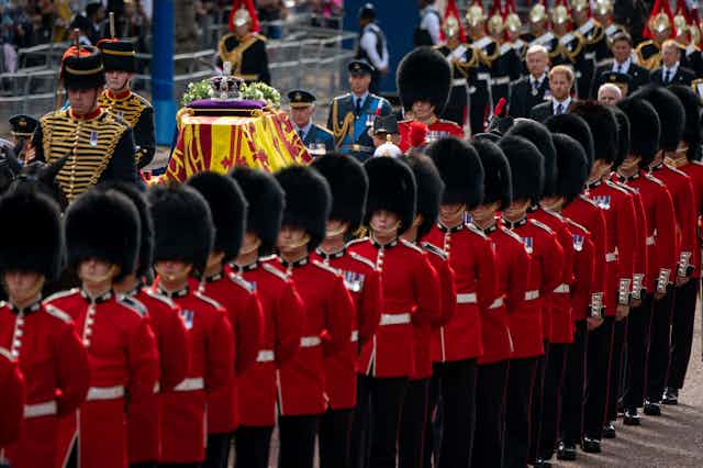 A line of guards in red coats and black fur hats escorts a coffin draped in a yellow and red standards and topped with flowers and a crown.