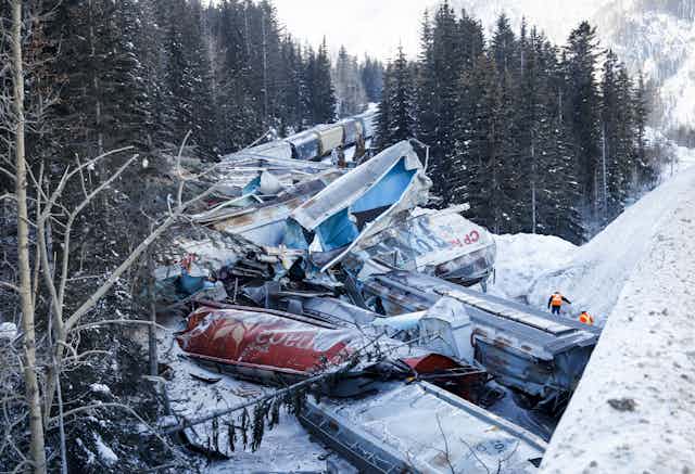 Crushed rail cars are seen in the snow amid pine trees.