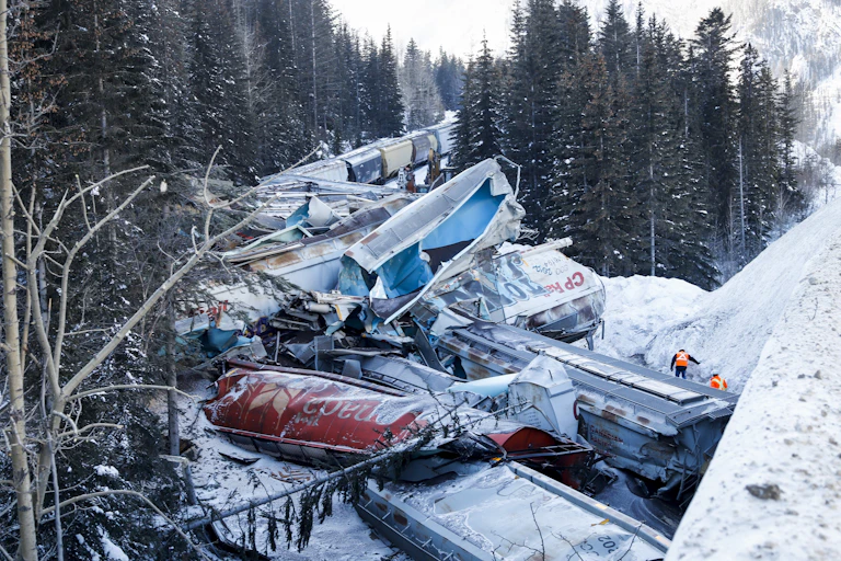 Crushed rail cars are seen in the snow amid pine trees.