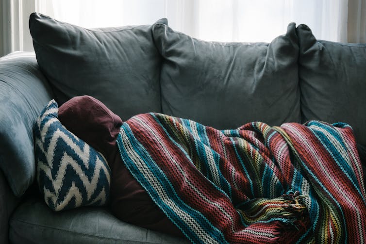 Person lays on couch in a hoodie, under a blanket.