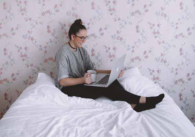 Woman sits on bed, talking on laptop, drinking tea