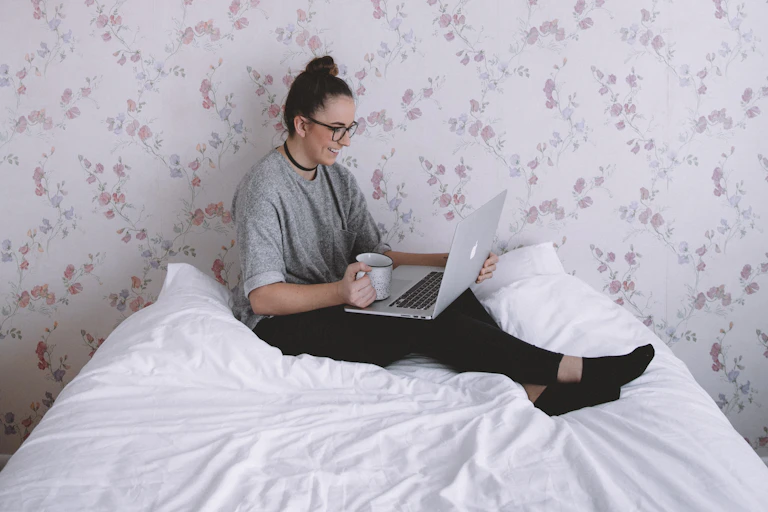 Woman sits on bed, talking on laptop, drinking tea