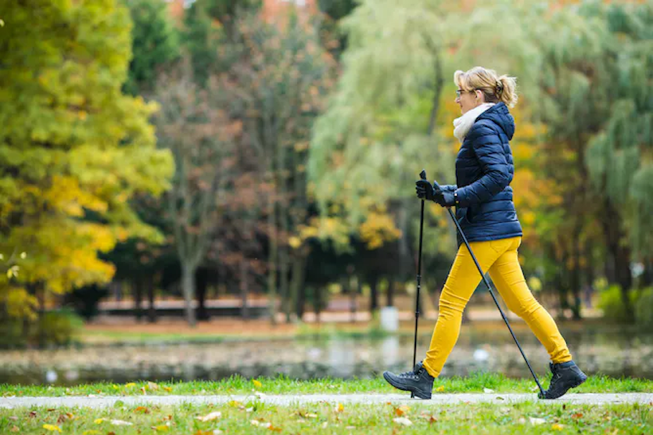 Seorang wanita yang mengenakan jaket musim dingin menggunakan tongkat berjalan Nordik saat berjalan melalui taman.