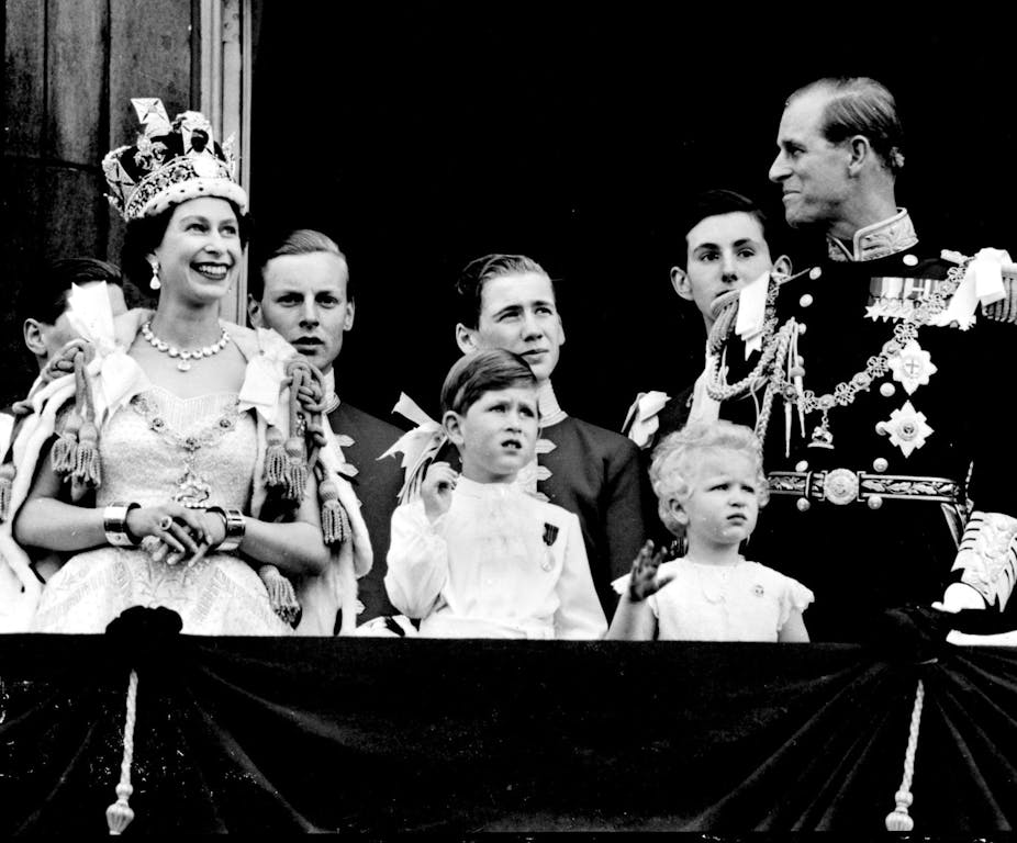 Black and white archive photo of a young Queen Elizabeth II, smiling on the balcony at Buckingham Palace wearing her coronation crown and robes, alongside her family