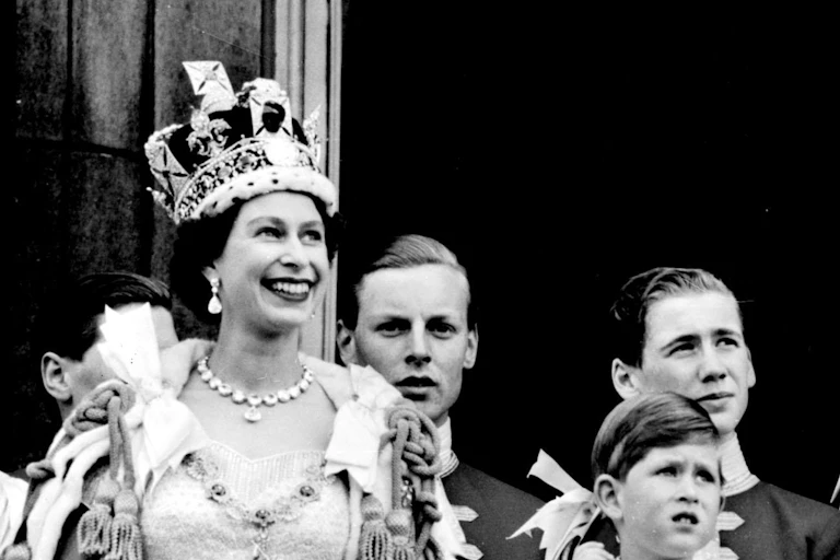 Black and white archive photo of a young Queen Elizabeth II, smiling on the balcony at Buckingham Palace wearing her coronation crown and robes, alongside her family