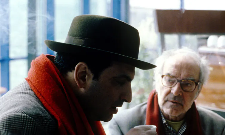 An old man in glasses talking to a younger man in a hat.
