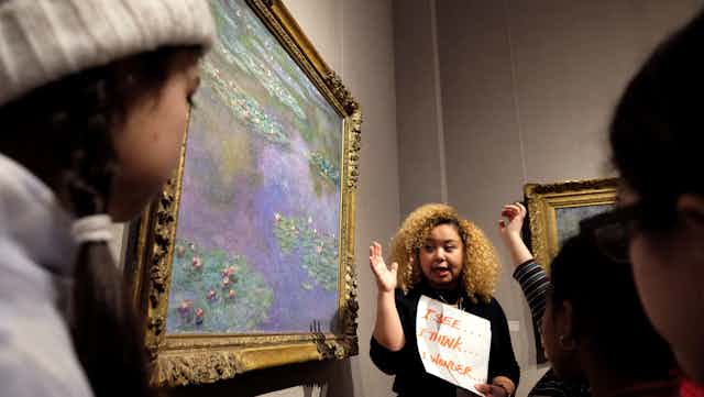 A woman stands next to a watercolor painting while addressing museum visitors.
