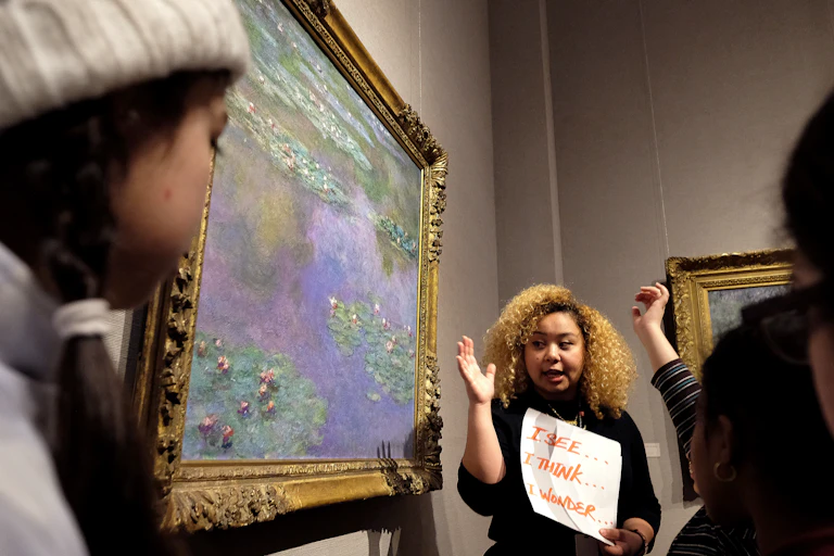 A woman stands next to a watercolor painting while addressing museum visitors.