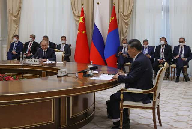 Russian President Vladimir Putin and Chinese President Xi Jinping sit at a roiund table while their aides watch from the side.