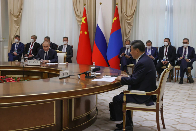 Russian President Vladimir Putin and Chinese President Xi Jinping sit at a roiund table while their aides watch from the side.
