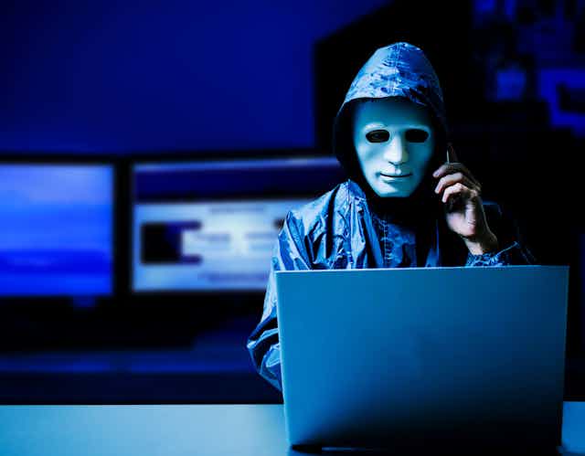 a man wearing a hooded jacket and with a sculpted mask over his face speaks on a phone while looking at a laptop in a dark room