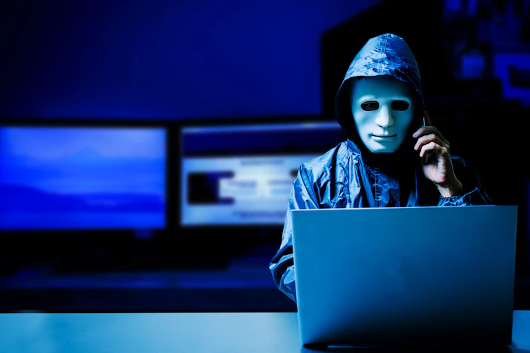 a man wearing a hooded jacket and with a sculpted mask over his face speaks on a phone while looking at a laptop in a dark room