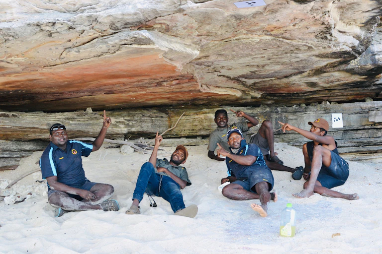 five men lie on sand pointing at rock overhang