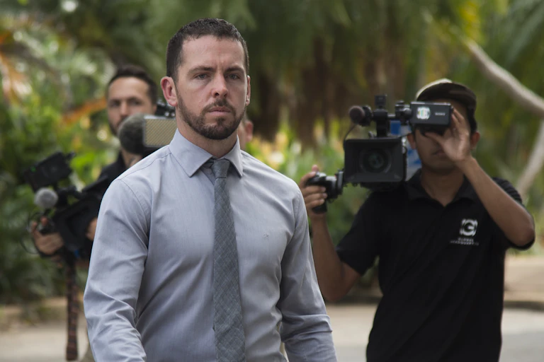 Zachary Rolfe, a white man in a shirt and tie is walking while video cameras are pointed at him.