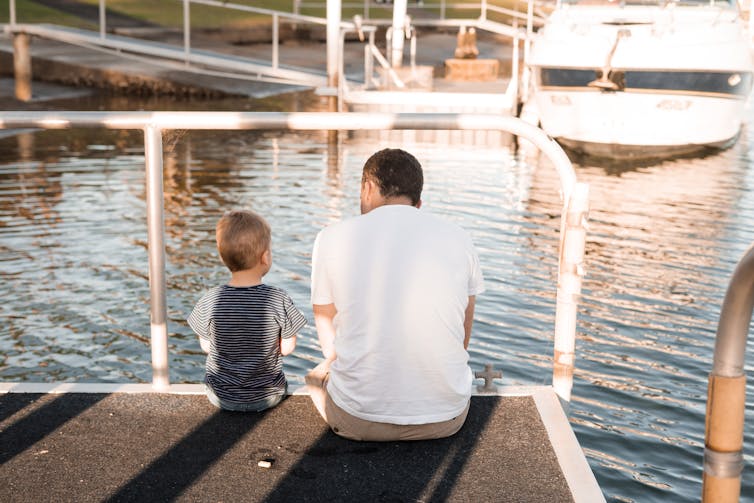 A father and son sit on a pier and talk.