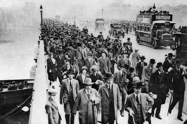 An archival photograph in black and white of a queue of people in hats and suits on a bridge over a river. 