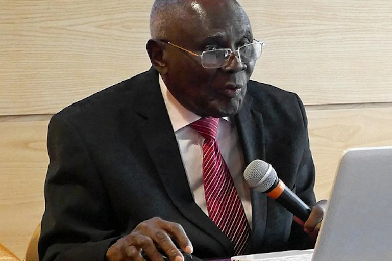 An elderly man in a suit and tie, sitting in front of a computer and holding a microphone