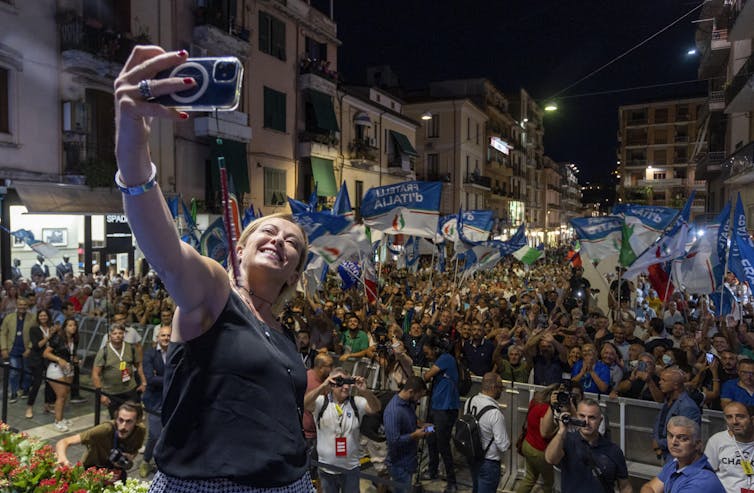 Giorgia Meloni takes a selfie at a political rally with a huge crowd behind her.