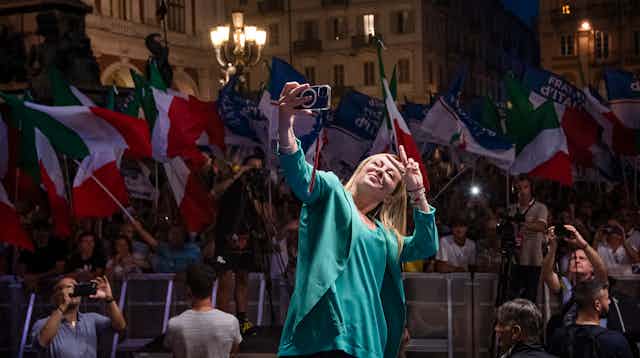Giorgia Meloni dressed in a green jacket and short flashes a 'v' sign while taking a selfie in front of supporters waving Italian flags.