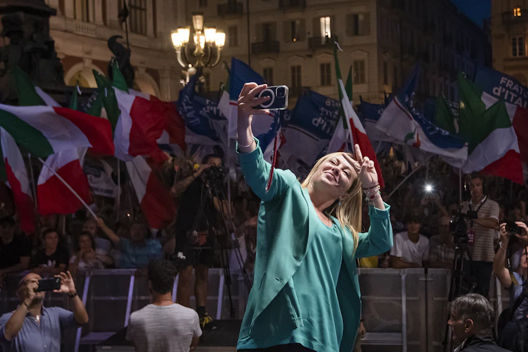 Giorgia Meloni dressed in a green jacket and short flashes a 'v' sign while taking a selfie in front of supporters waving Italian flags.