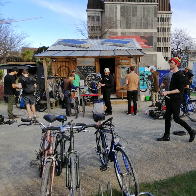 In the foreground three bikes on a stand. In the background, a recycled tiny workshop with people repairing bikes