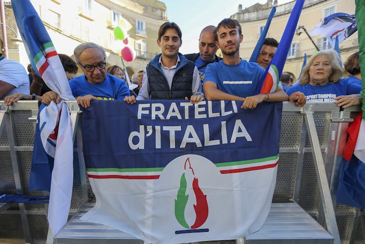 A group of men stand around a flag with 'Fratelli d'Italia' on it and a flame symbol below.
