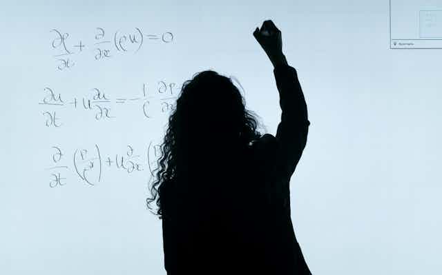 A black silhouette of a woman writing equations on a whiteboard
