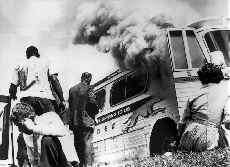 Black and white photograph of people standing and sitting outside of a burning bus.