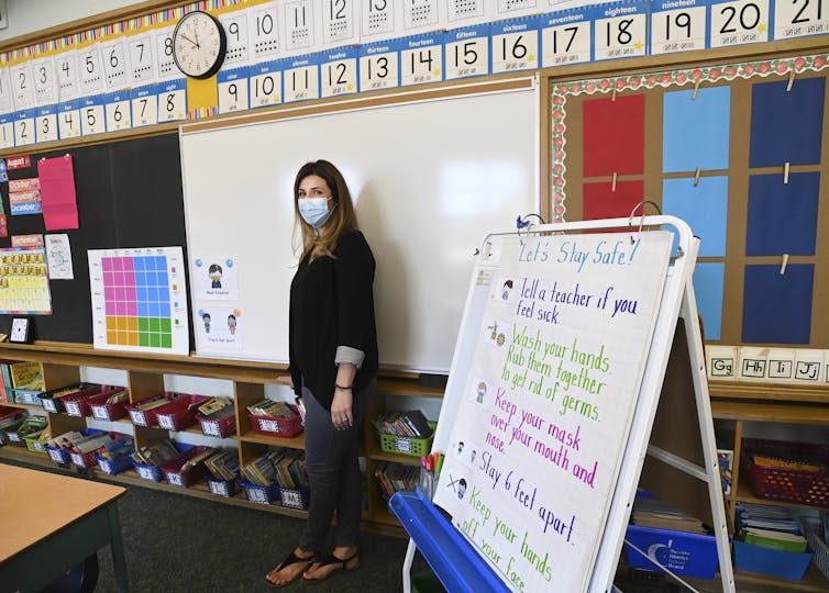 A teacher wears a mask next to a list of pandemic rules in the classroom about keeping one's mask on and washing hands.