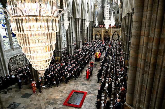 Rows of people in black clothing stand in a huge cathedral as people carry out a coffin draped in a flag.