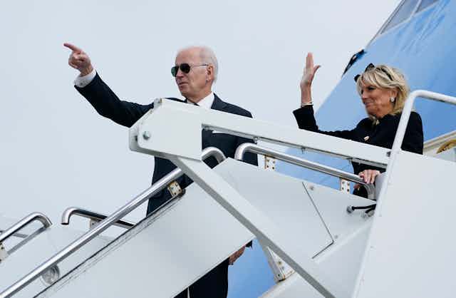 An older man in a black suit and sunglasses points from the stairs of an airplane; a blond woman behind him waves. 