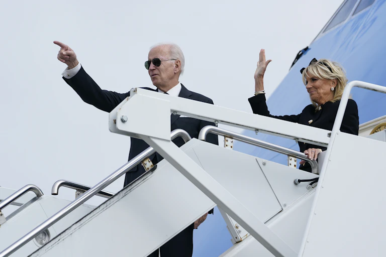 An older man in a black suit and sunglasses points from the stairs of an airplane; a blond woman behind him waves.