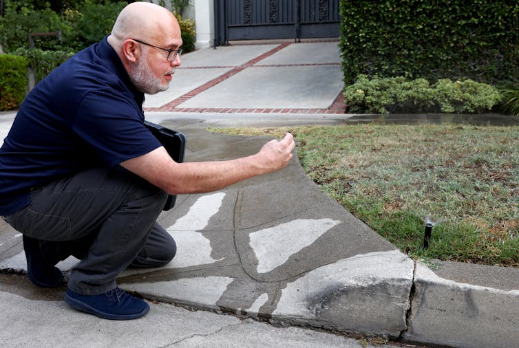 A man in a blue uniform stoops at the end of a driveway observing water flowing into the street.