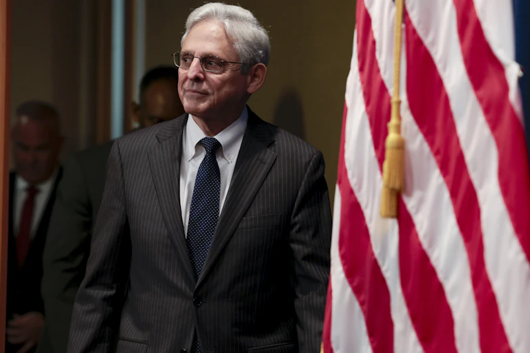 A middle-aged white man dressed in a business suit appears on stage standing next to an American flag.