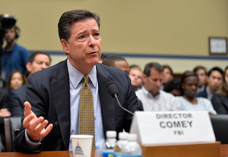 A white man dressed in a business suit gestures with his hand as he answers a questions while sitting behind a table.