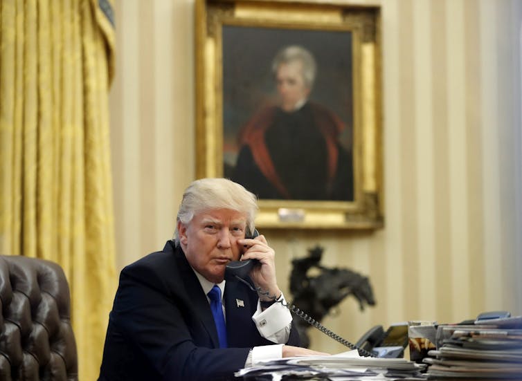 A grey-haired man talks on the phone with a 1800s-era portrait of a man hanging on the wall behind him.