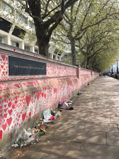 A concrete wall covered in drawings of thousands of red hearts in different sizes, with bouquets of flowers lining it on the ground.