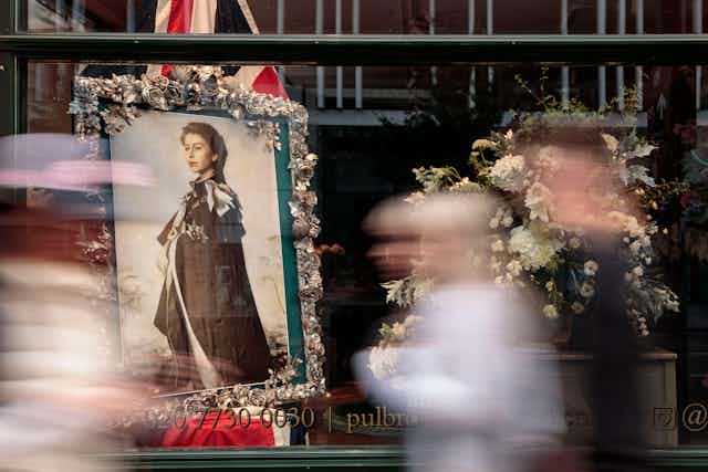A photograph of a photograph of the Queen in a shop window with people passing fast, their silhouettes all blurred.